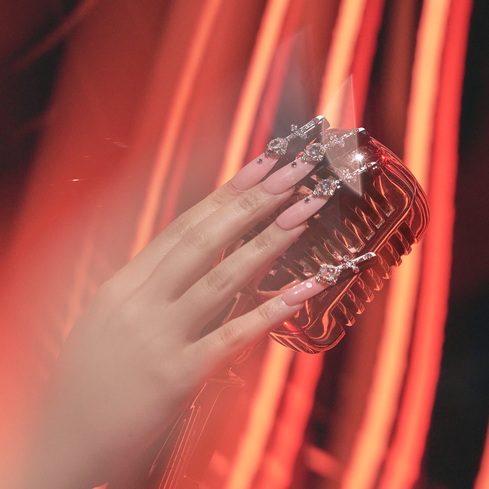 Hand showcasing MiuNail FRAME collection Divine Plea press-on nails with bold crystal details, in dramatic orange lighting.