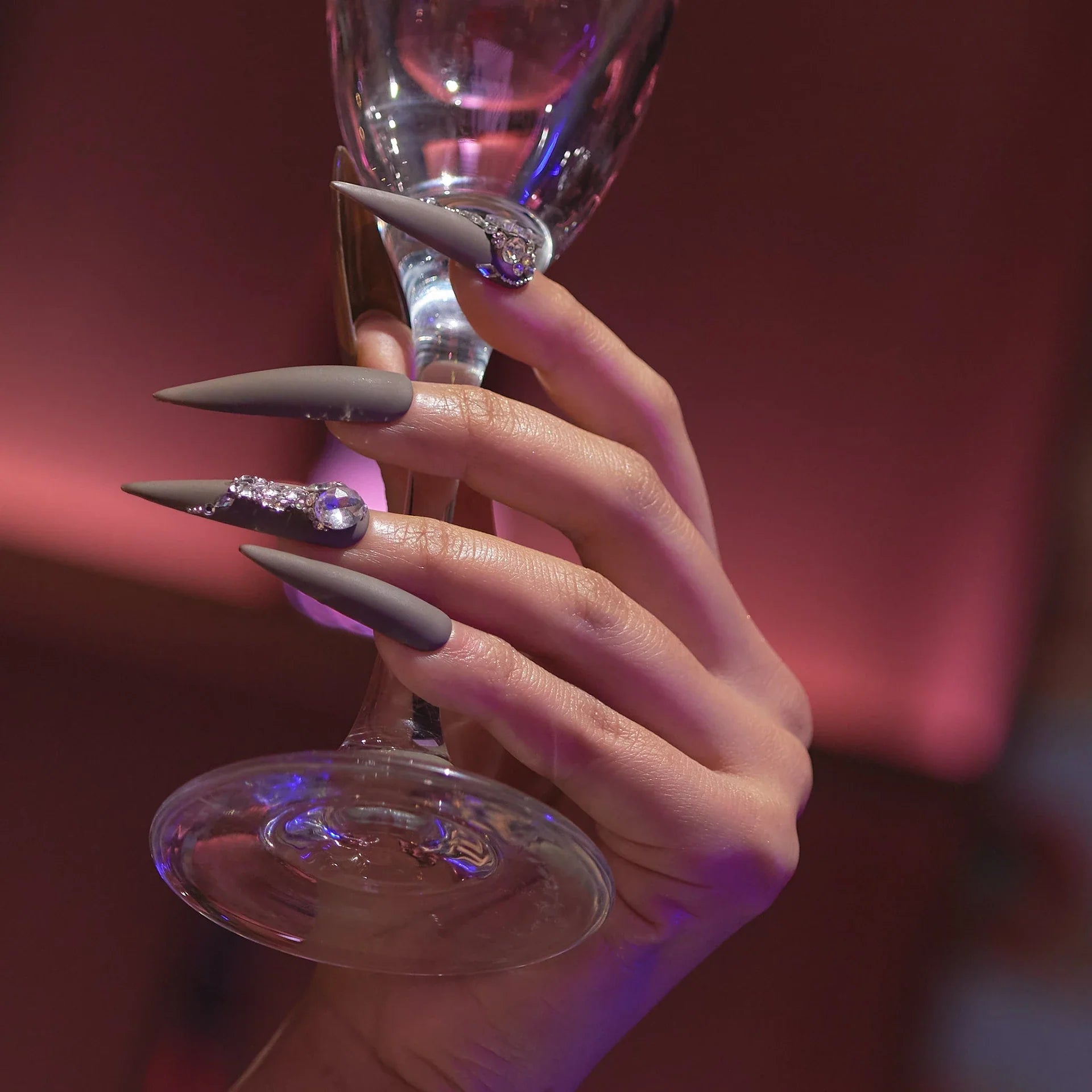 Gala collection press-on nails, long stiletto in matte gray with rhinestone accents, holding wine glass.