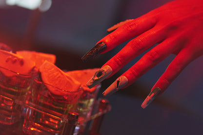 Hand with MiuNail Siren Hex press-on nails in icy blue and white tones hovering over red-lit shot glasses with lime wedges.