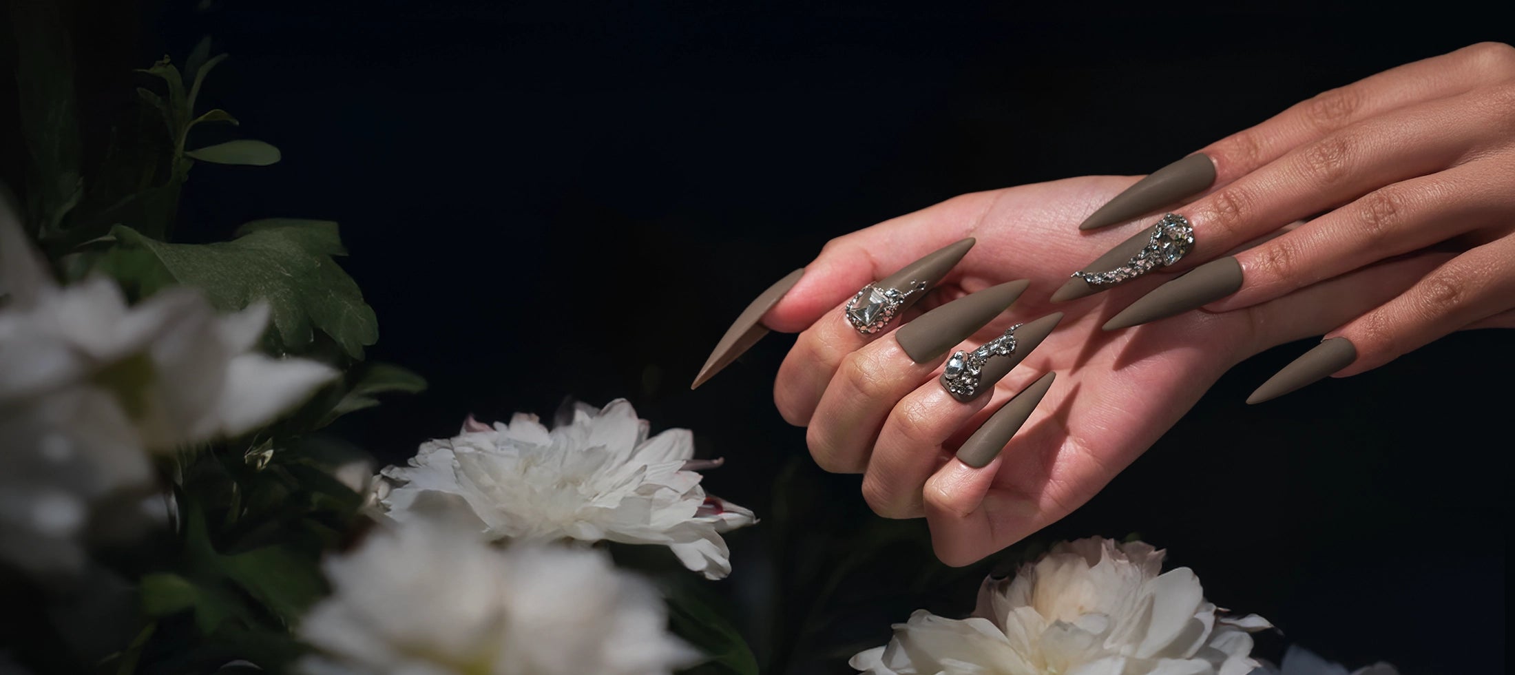 Hand wearing MiuNail “Desert Halo” long matte press-on nails with crystal rhinestone accents, shown against a soft floral background.