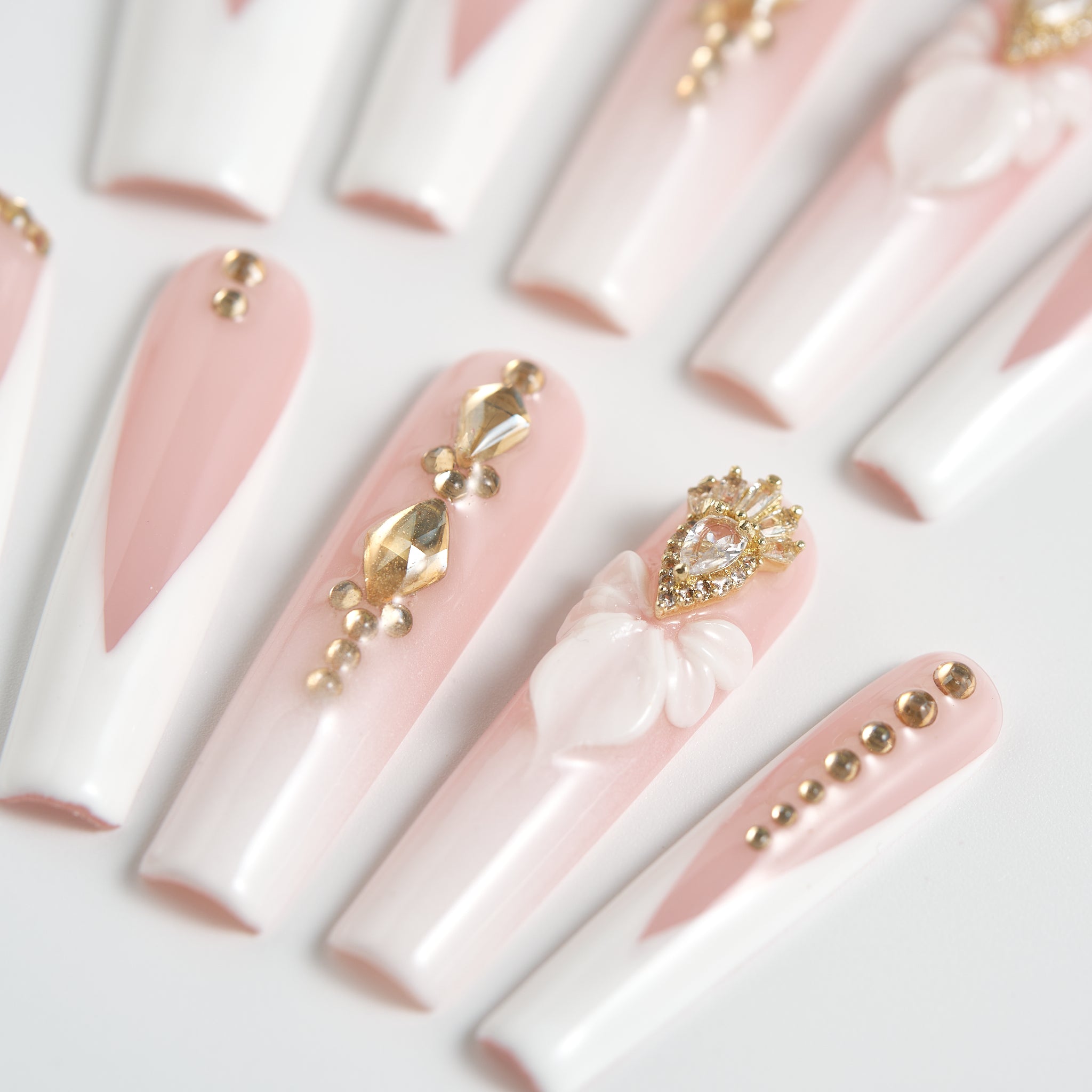 angled view of long square nails in blush pink and white with gold rhinestones and 3D bow accents on a white background
