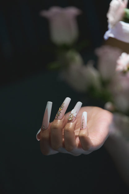 close-up of long square nails in blush pink and white with gold embellishments and 3D bow shown against a blurred floral background