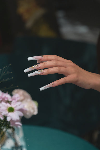 long square nails in blush pink and white with gold rhinestones and 3D bow shown on an extended hand near pink flowers on a blurred teal background