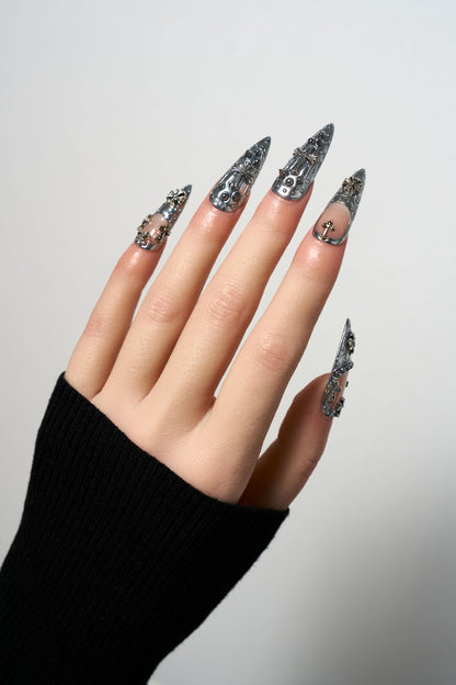 Chrome Reign reusable stiletto press-on nails with a high-shine silver metallic finish, featuring intricate gothic embossed patterns, 3D silver cross charms, and punk-style rivet details.