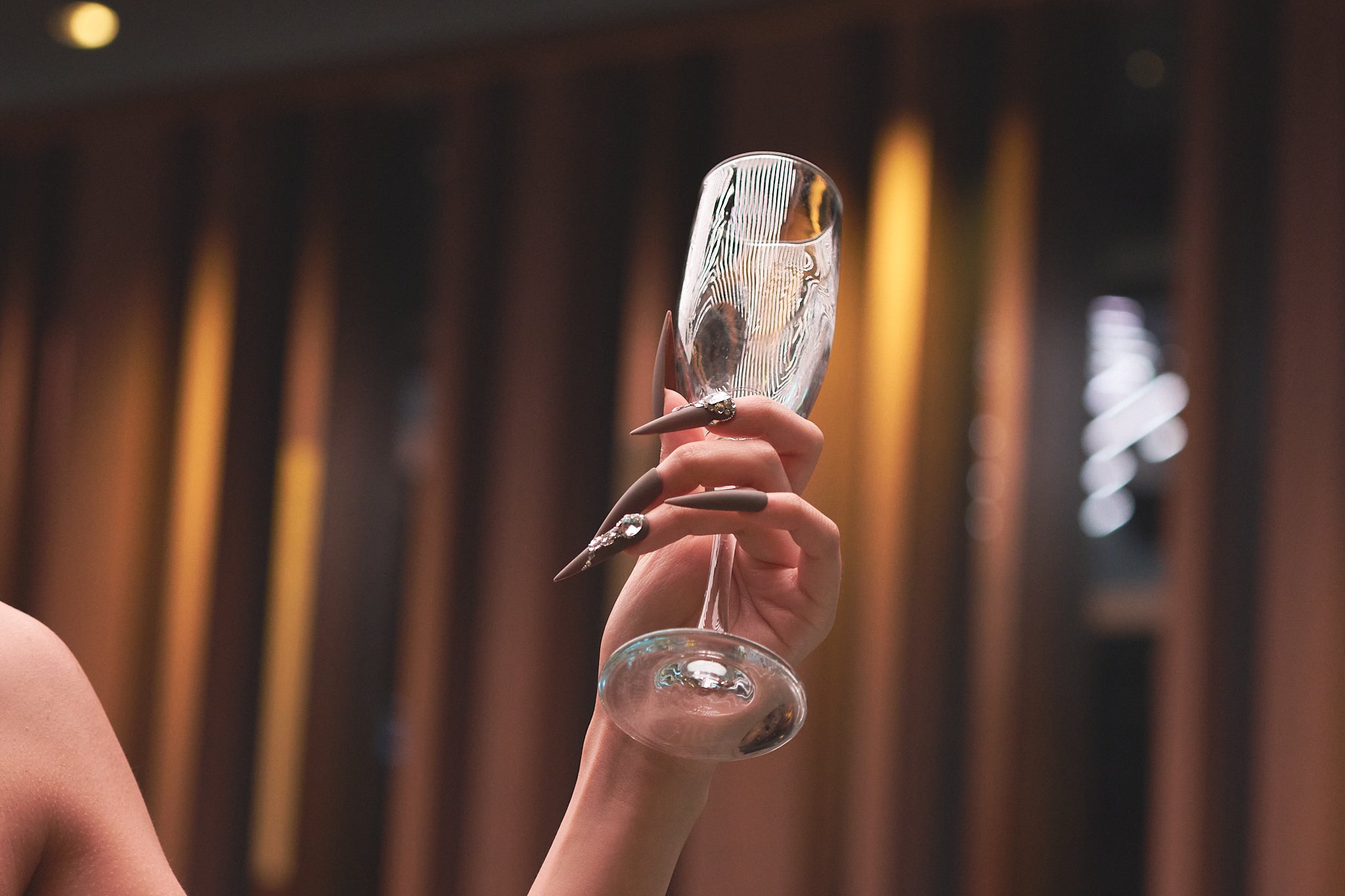 Hand wearing long stiletto ‘Desert Halo’ press-on nails holding a champagne flute against a modern blurred backdrop.
