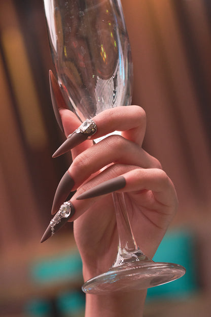 Close-up of long stiletto ‘Desert Halo’ press-on nails with crystal nail art, holding a champagne flute.
