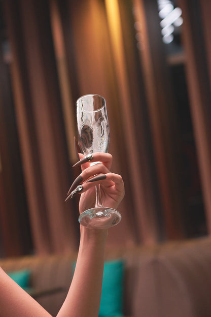 Angled view of hand with long stiletto ‘Desert Halo’ press-on nails and crystal accents, holding a champagne flute.