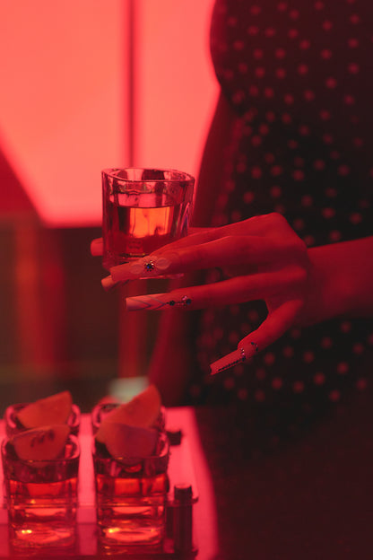 Hand with ‘Frost Waltz’ long square press-on nails holding a shot glass under red lighting at a bar, with tequila and lime in the foreground.