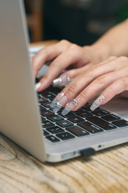 Close-up of hands wearing Gilded Heiress beige rhinestone press-on nails typing on a laptop keyboard.
