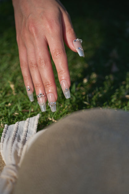Close-up of a hand with Gilded Heiress decorative rhinestone nails resting on a grassy outdoor background, with a beige hat partially visible.