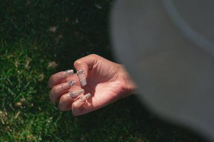 Close-up of a hand with Gilded Heiress decorative rhinestone nails outdoors on grass.