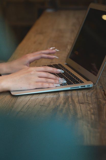 Hands with Gilded Heiress beige rhinestone press-on nails typing on a laptop at a wooden table.