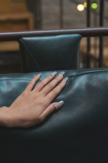 Hand with Gilded Heiress beige rhinestone press-on nails resting on a black leather chair.