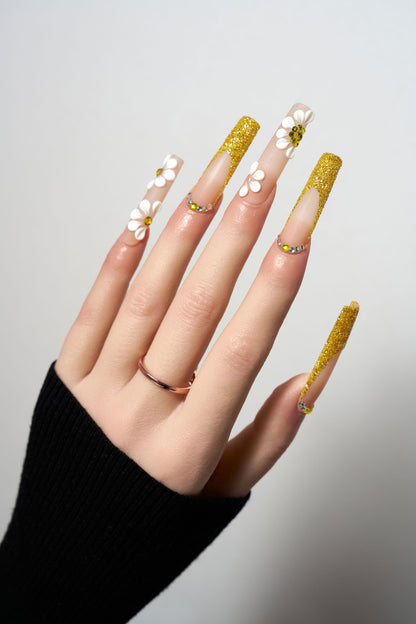 Golden Bloom reusable extra-long coffin press-on nails featuring gold glitter French tips, decorated with 3D white daisy flowers with amber centers and delicate crystal borders.
