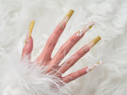Hand wearing ‘Golden Bloom’ long square press-on nails with gold glitter tips and 3D white daisy accents, posed over a white faux fur background.