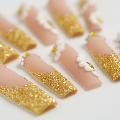 Depth-focused macro of ‘Golden Bloom’ long square press-on nails featuring gold glitter, white 3D daisies, and gemstone detailing on a beige base.