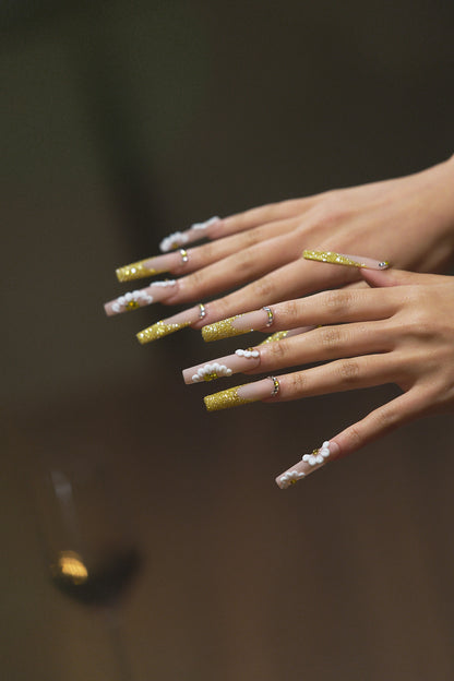 long square nails in gold glitter and 3D white florals shown on both hands over a blurred background