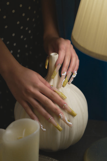 long square nails in gold glitter and white 3D flowers touching a white pumpkin on dark background