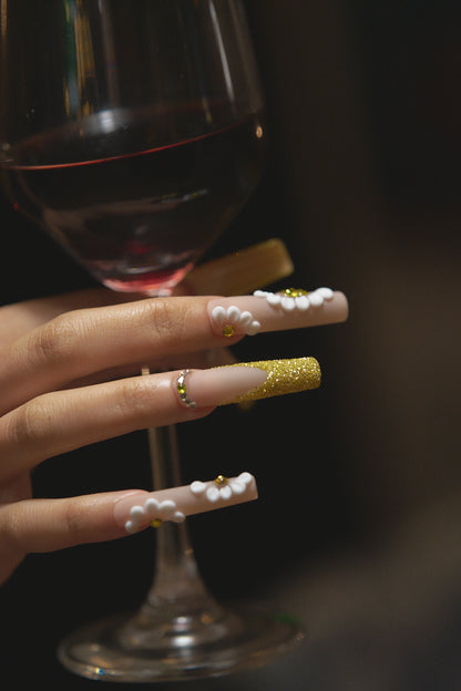 close-up of long square nails in gold glitter with 3D white florals holding a red wine glass on dark background