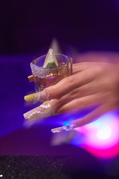 long square nails in gold glitter with 3D white florals holding a salt-rimmed shot glass with lime against purple lighting