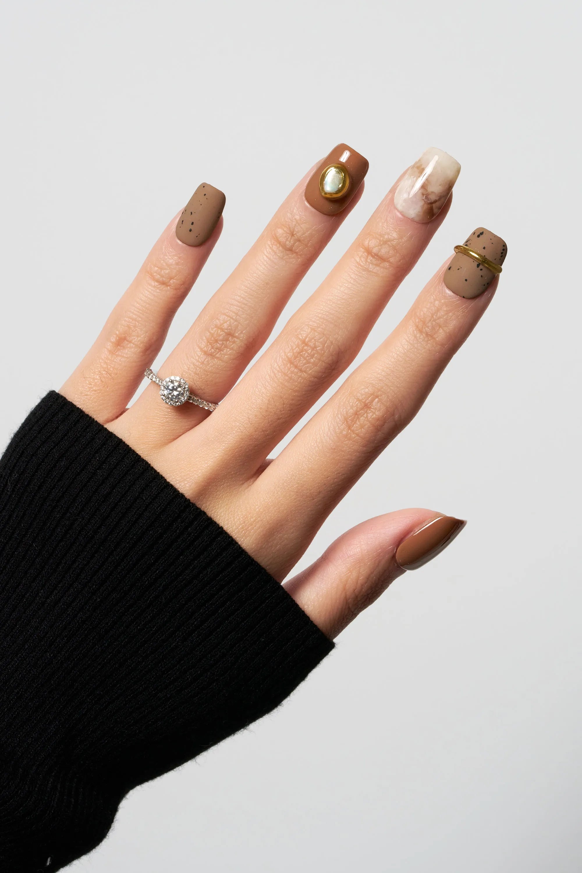 Golden Dune press-on nails featuring a mix of matte earthy brown with black speckles, cream marble swirl design, and a unique 3D gold ring accent on a square nail shape.