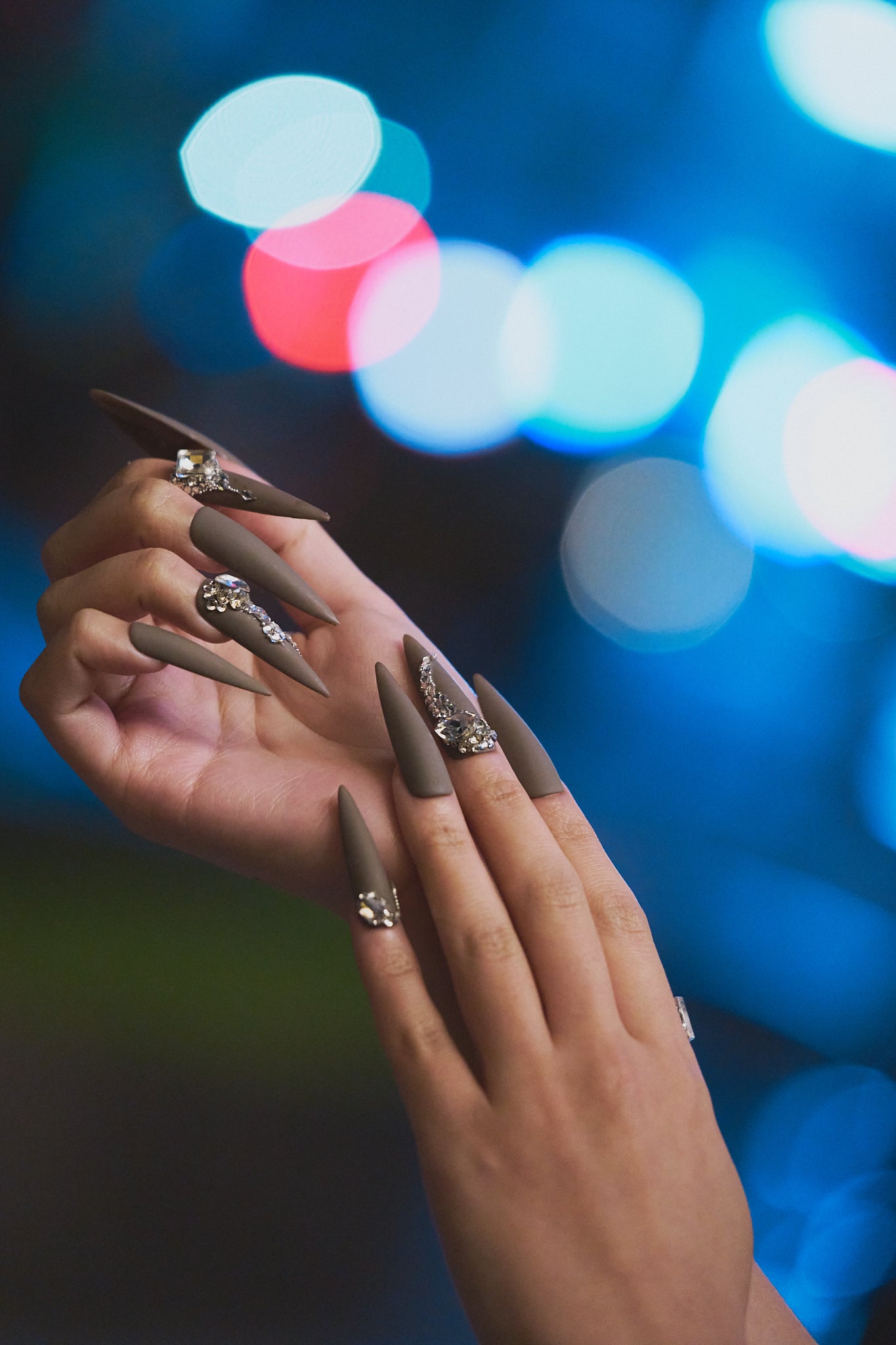 Hands wearing long stiletto brown press-on nails with rhinestone details, shown against a colorful bokeh night background.