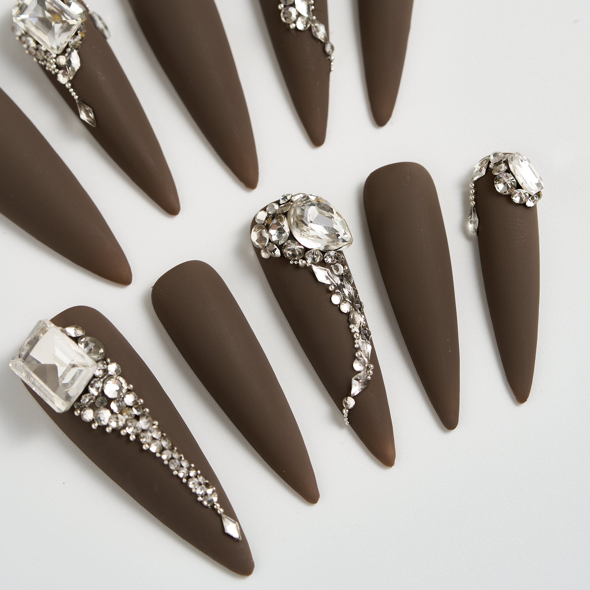Close-up of long stiletto matte brown press-on nails with crystal and rhinestone embellishments, displayed on a white background.