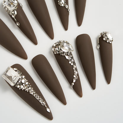 Close-up of long stiletto matte brown press-on nails with crystal and rhinestone embellishments, displayed on a white background.