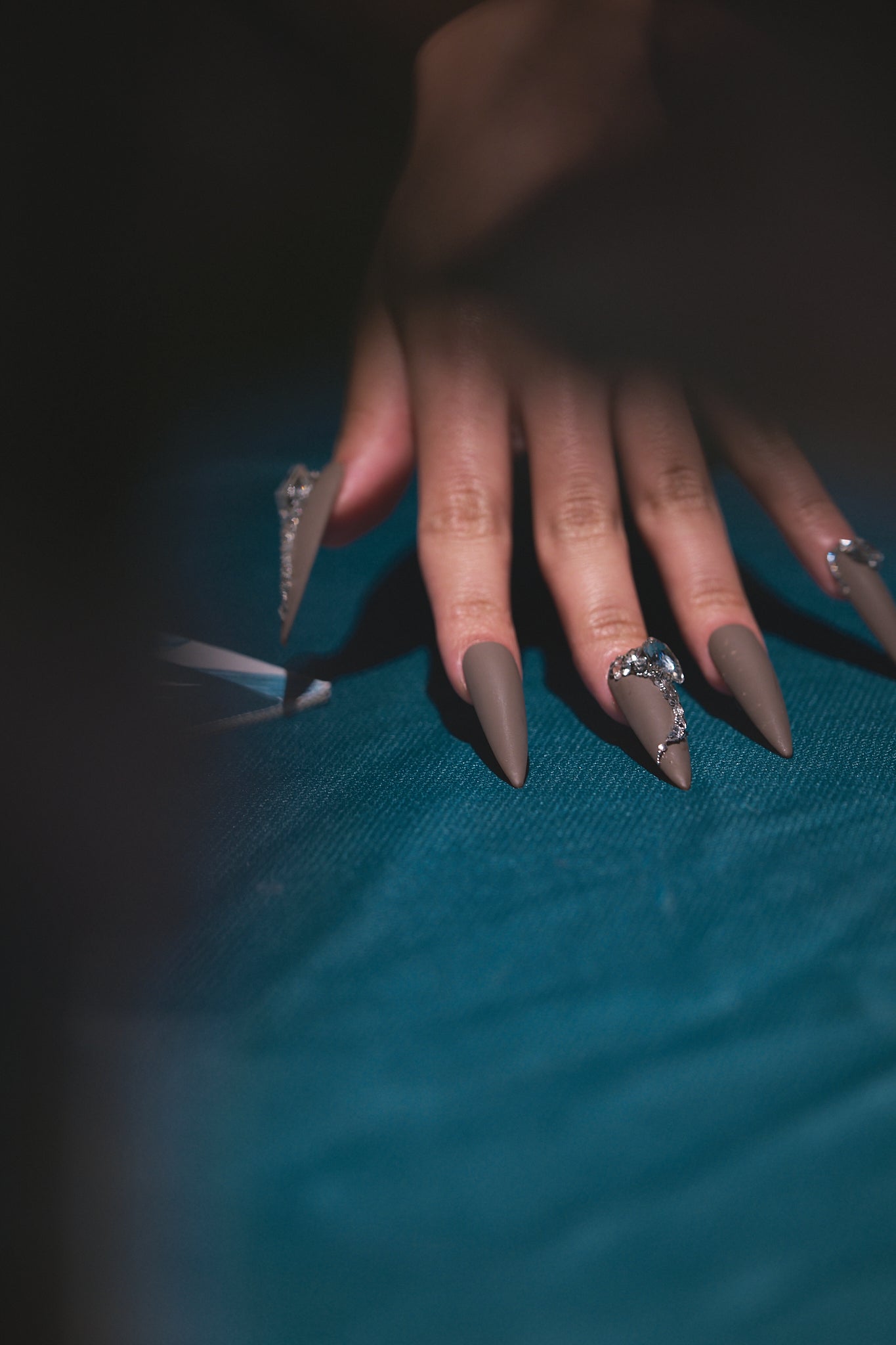 Close-up of a hand with long matte brown stiletto nails and rhinestone accents resting on a dark blue fabric.