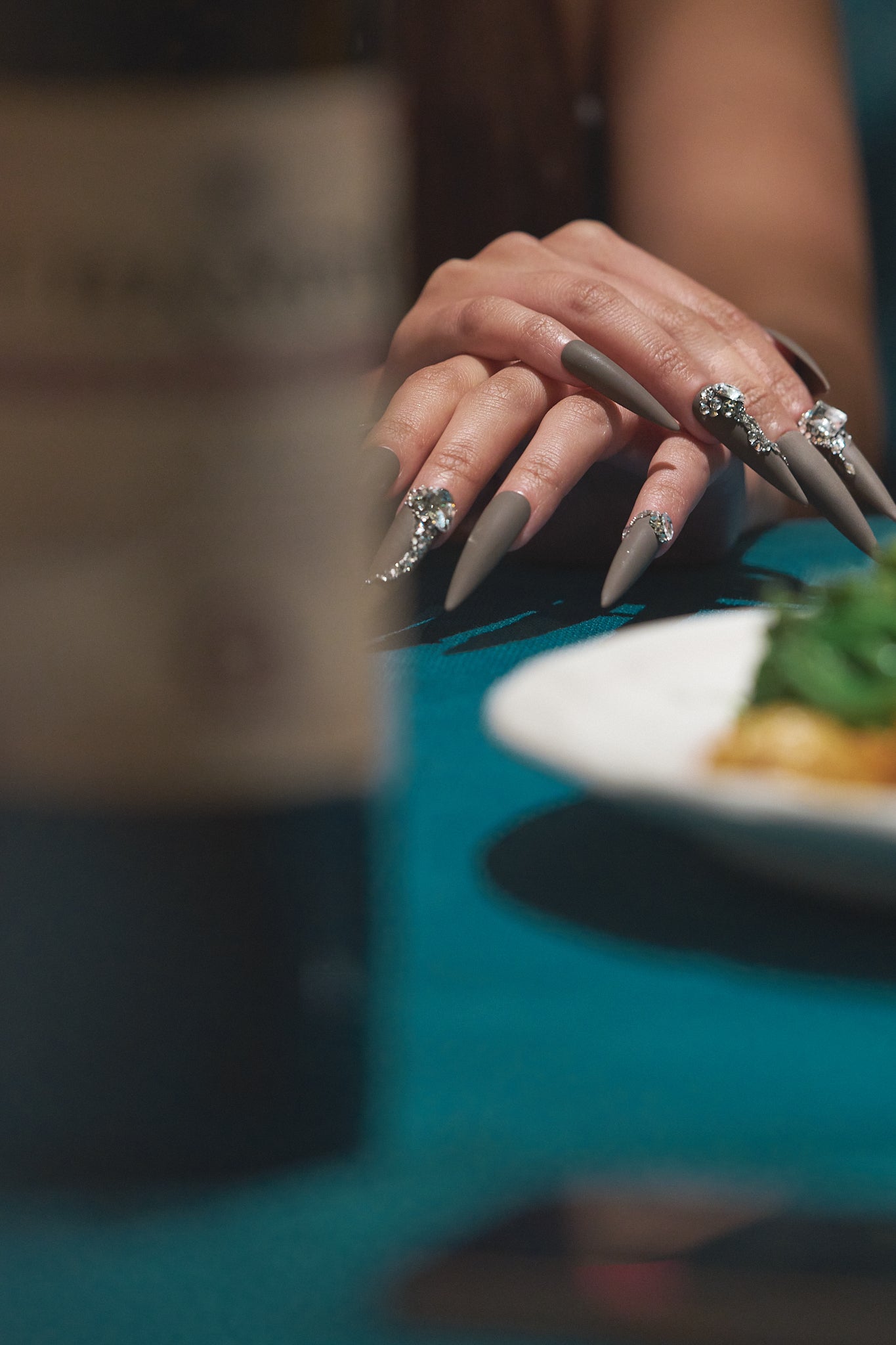 Close-up of hands wearing long stiletto brown press-on nails with rhinestones, holding a fork over a dinner table.