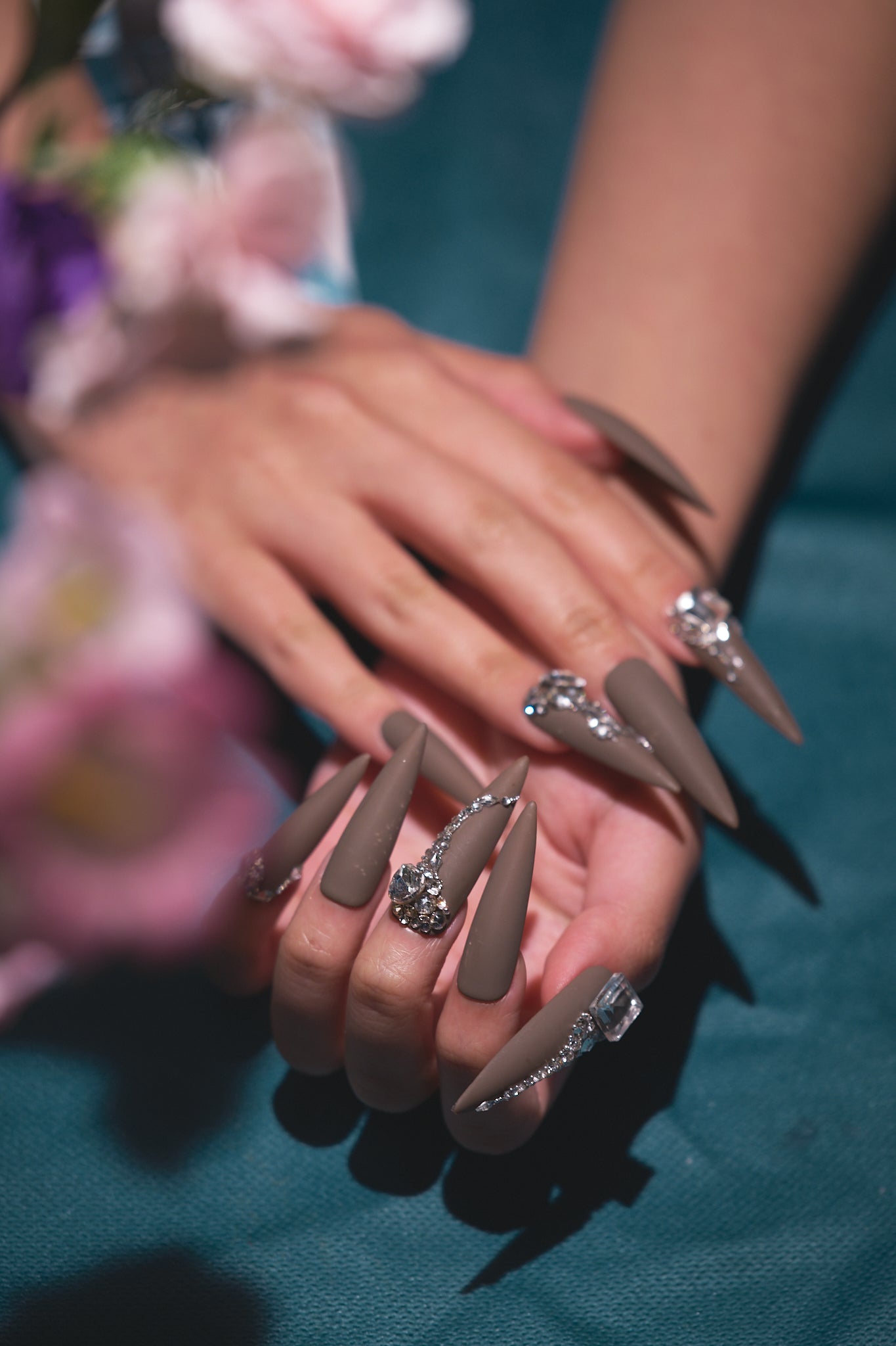 Two hands with long brown matte stiletto nails adorned with rhinestones, resting on a teal surface with blurred floral background.
