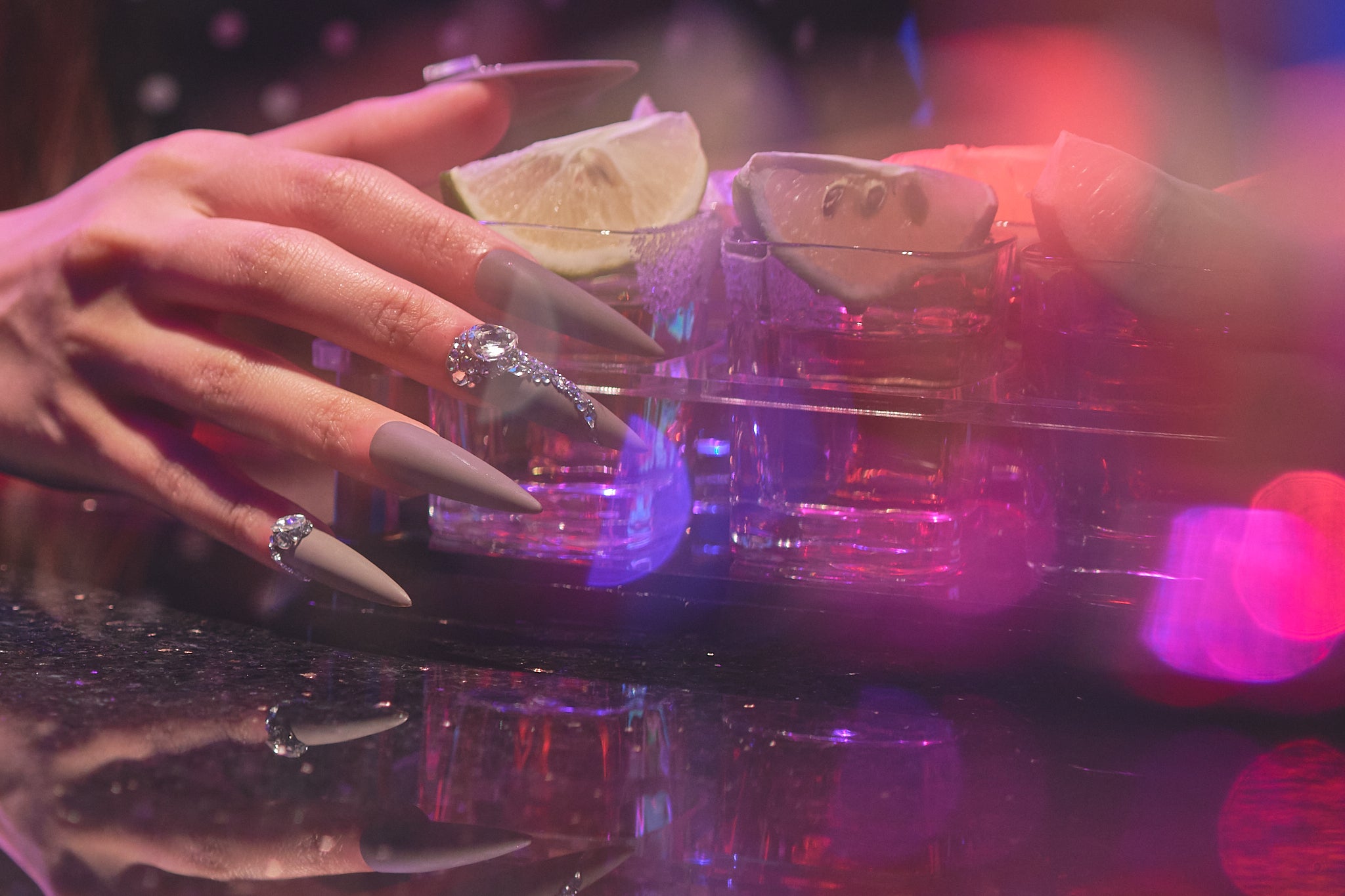 Close-up of a hand with long brown stiletto nails and rhinestone details holding a glass of tequila with lime wedges, under pink and purple ambient lighting.