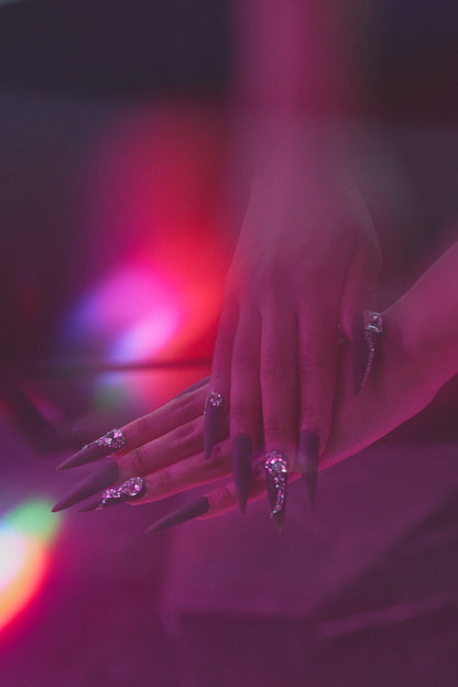 Close-up of hands with long brown stiletto nails and rhinestone accents under soft, dreamy purple and red lighting.