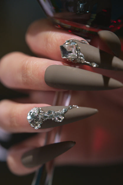 Close-up of hand wearing long stiletto matte brown press-on nails with rhinestone decorations, holding a wine glass.