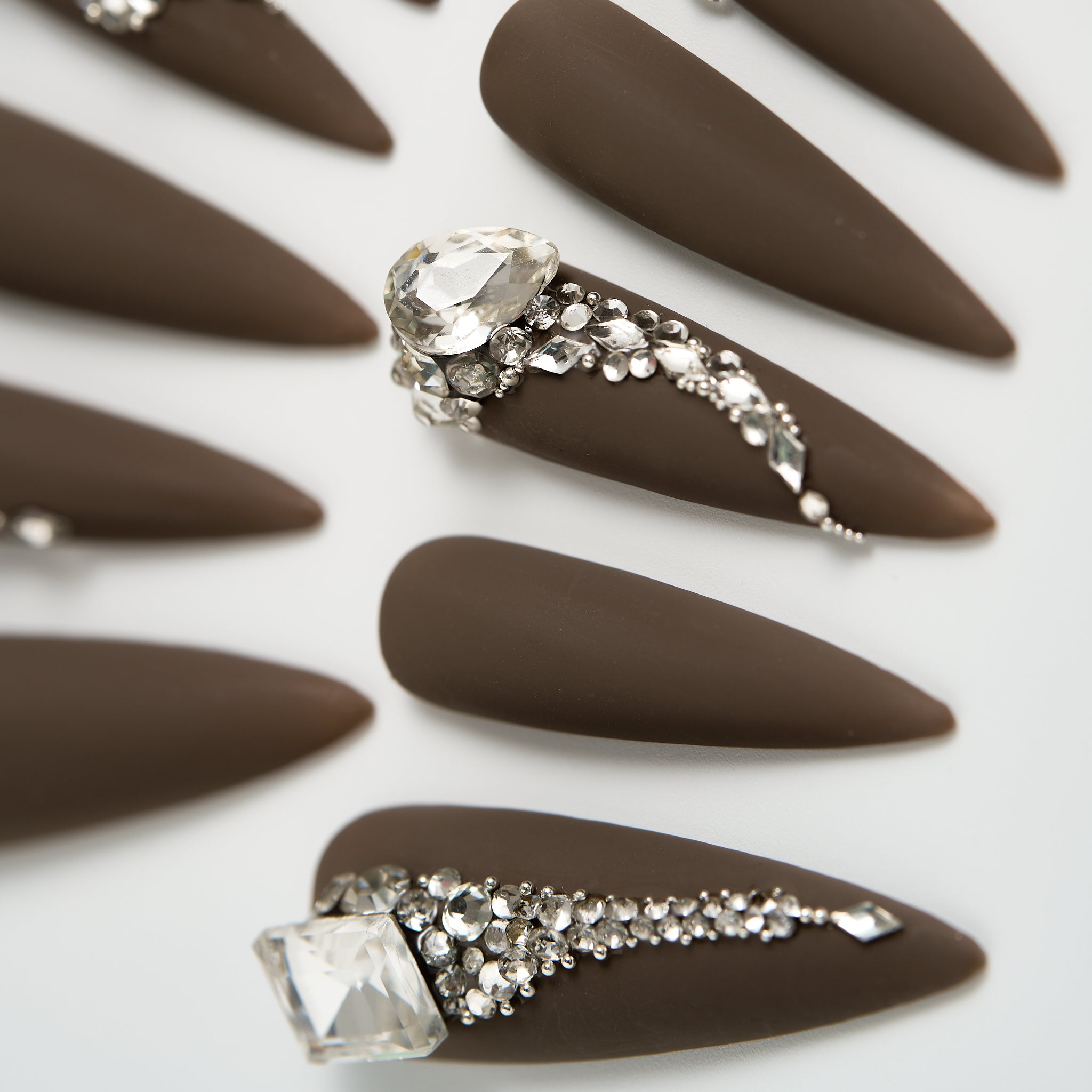 Detail shot of long stiletto matte brown press-on nails with large crystal rhinestone accents on a white background.