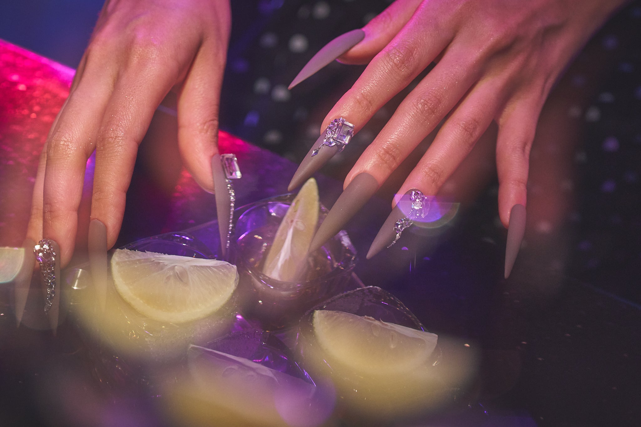 Close-up of hands with long brown stiletto nails and rhinestone accents over glasses of tequila with lime wedges, set under vibrant purple-pink lighting.