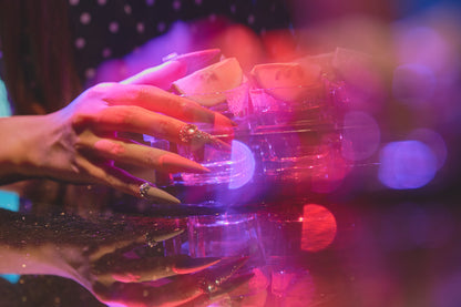 Side view of hands with long brown stiletto nails and rhinestone accents holding a glass of tequila with lime, set against vibrant purple and pink bokeh lighting with mirrored reflections.
