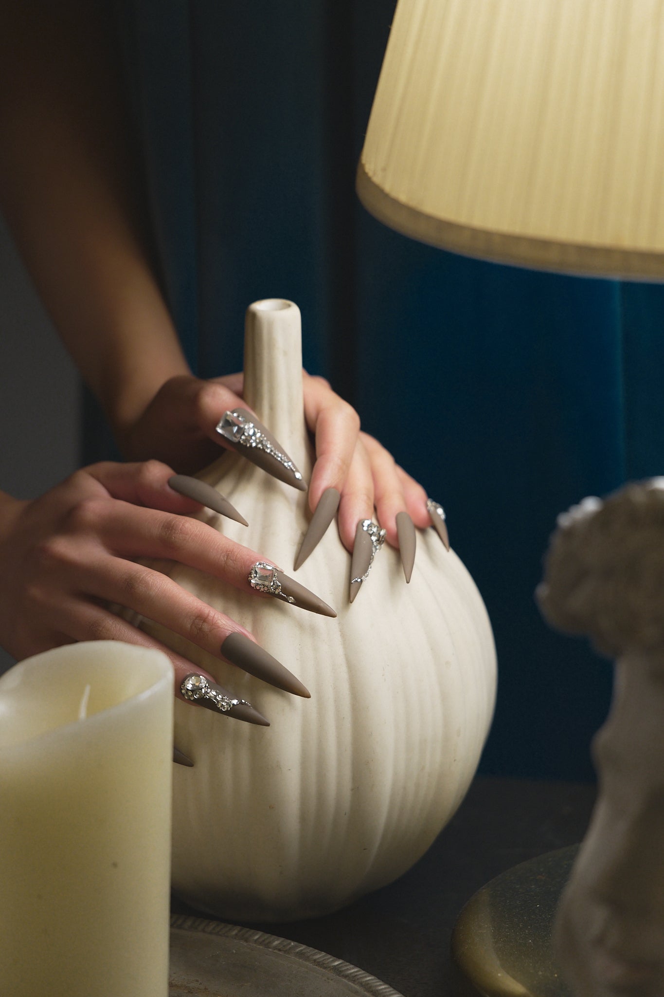 Hands with long brown stiletto nails decorated with rhinestones, gently resting on a ceramic vase beside candles and a lamp.