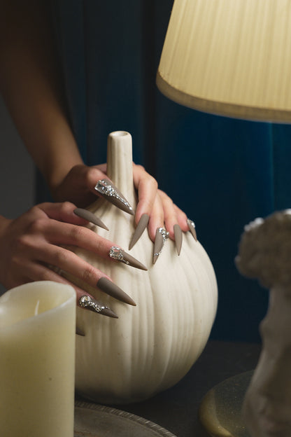 Hands with long brown stiletto nails decorated with rhinestones, gently resting on a ceramic vase beside candles and a lamp.
