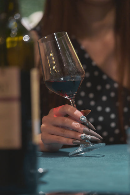 Hand wearing long brown stiletto nails with rhinestone accents, holding a wine glass filled with red wine, blurred background.