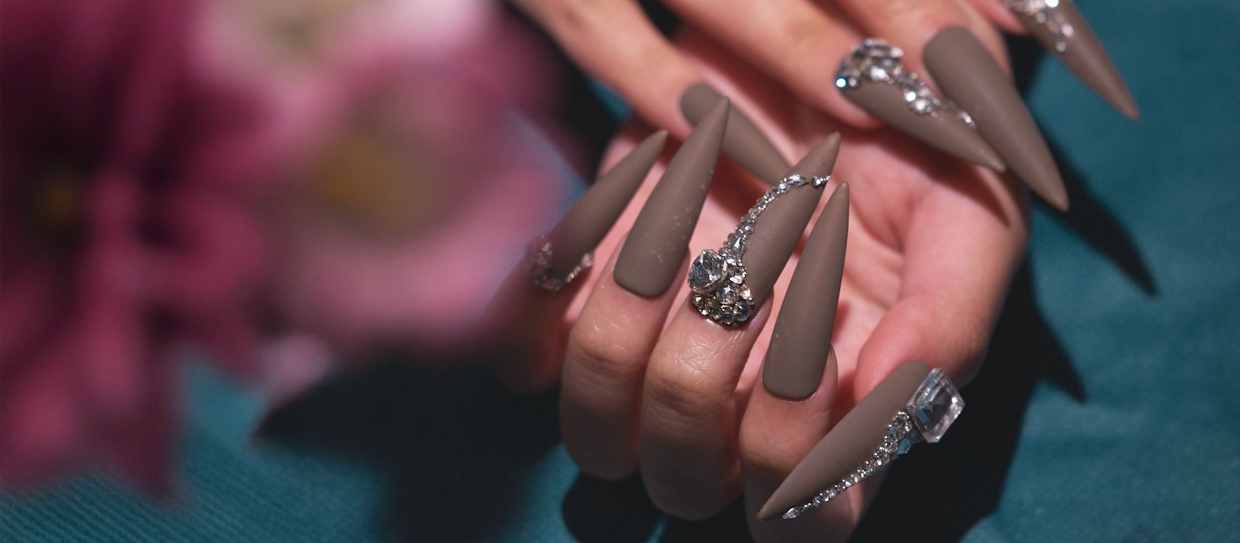 Hand wearing MiuNail “Desert Halo” long matte press-on nails with crystal rhinestone accents, shown against a soft floral background.