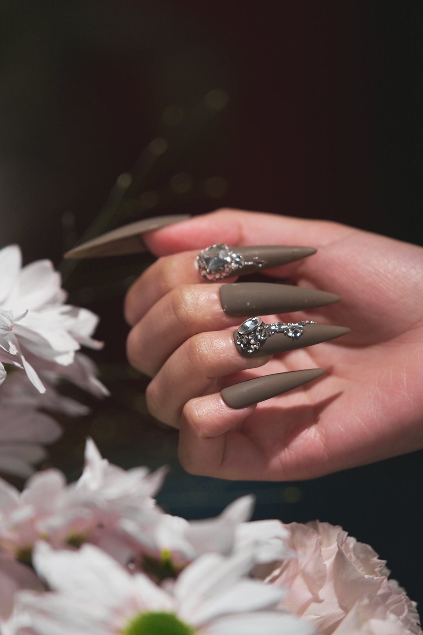 Close-up of a hand with matte brown rhinestone nails holding pale pink flowers in soft daylight.