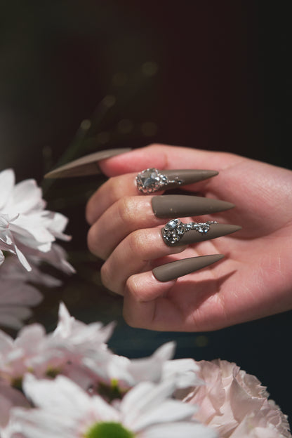 Close-up of a hand with matte brown rhinestone nails holding pale pink flowers in soft daylight.