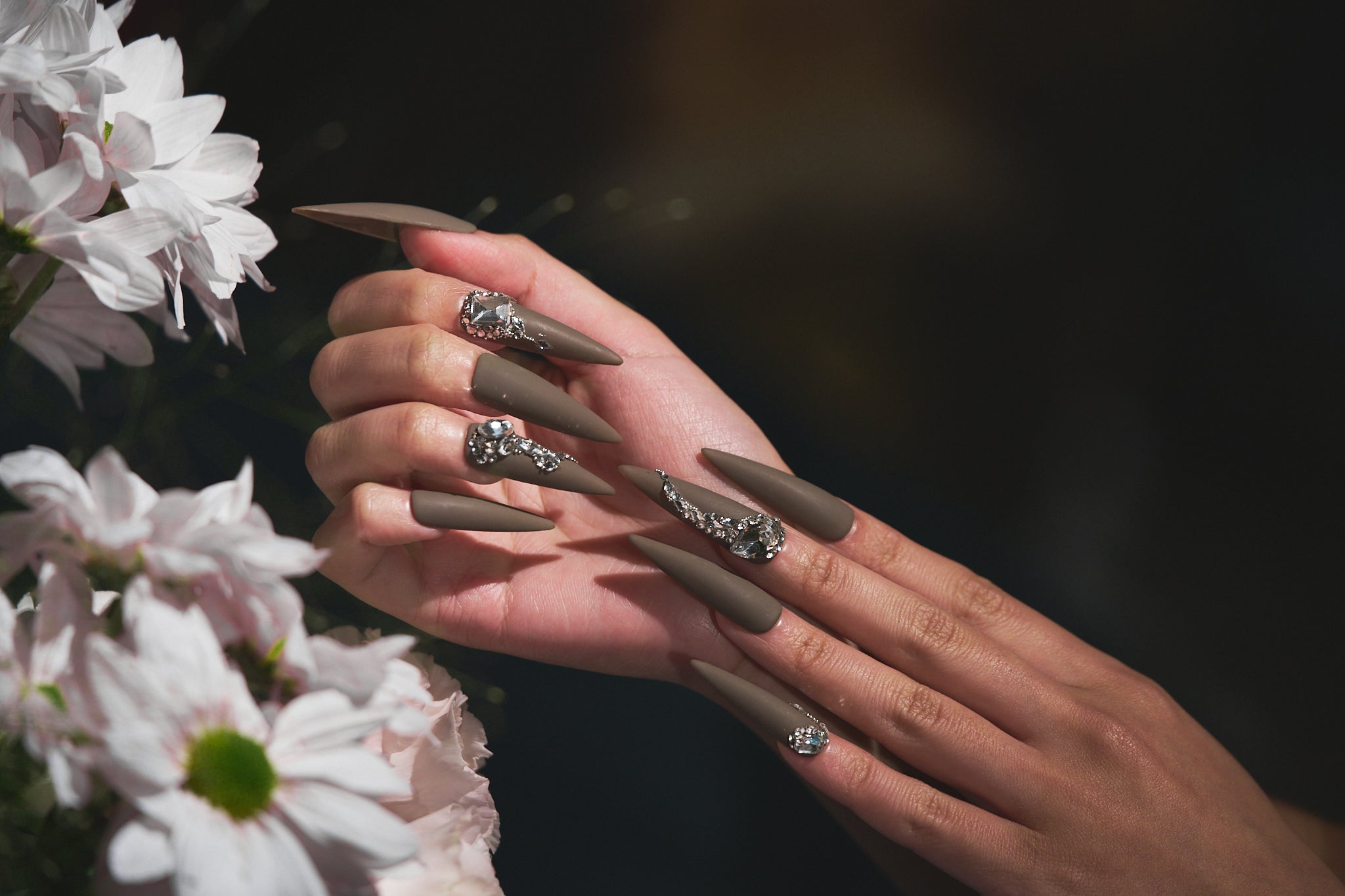 Hands with long matte brown stiletto nails and rhinestone accents, holding white flowers in soft natural light.