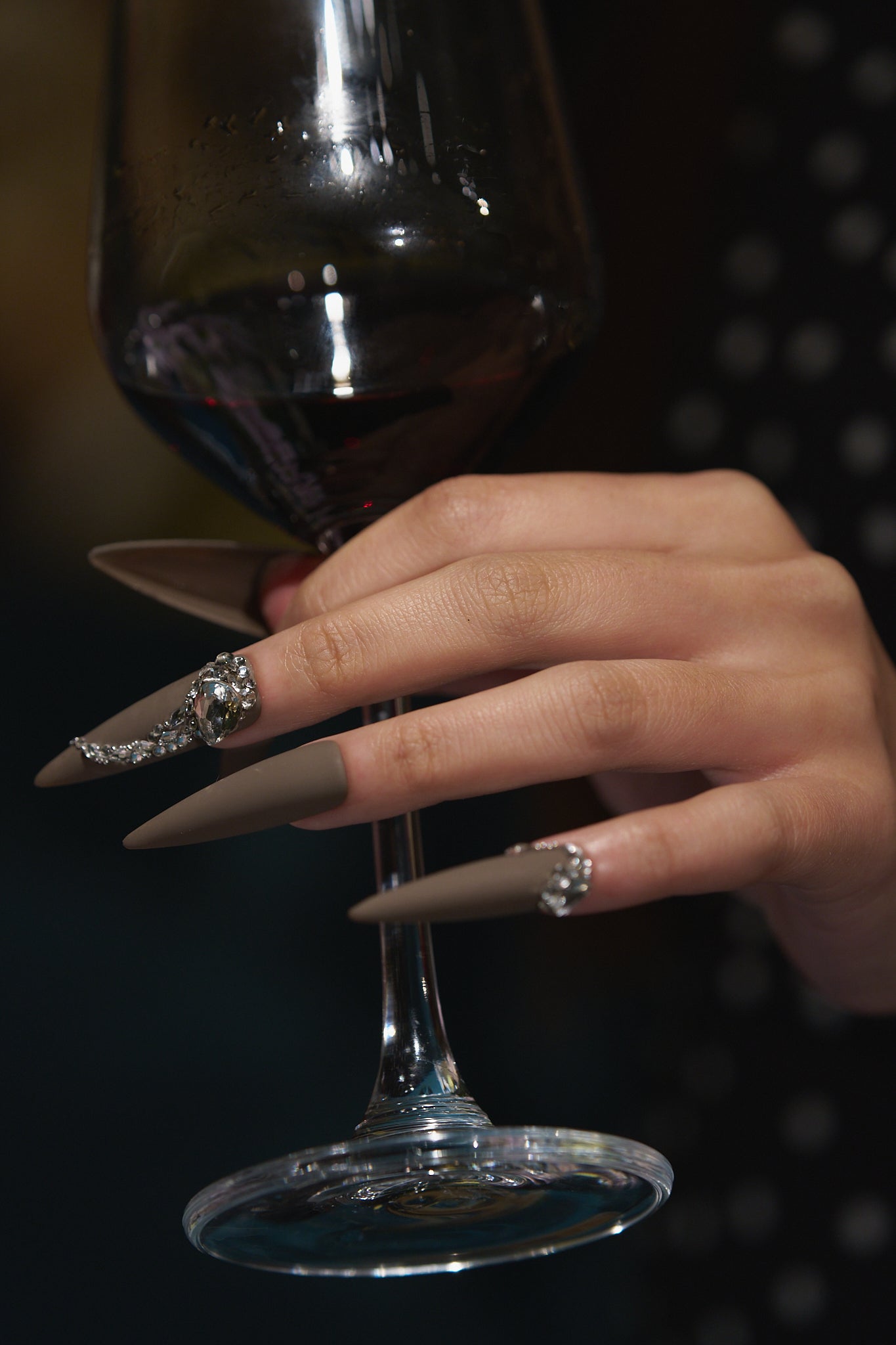 Close-up of a hand with long brown stiletto nails decorated with rhinestones, holding a glass of red wine against a dark background.