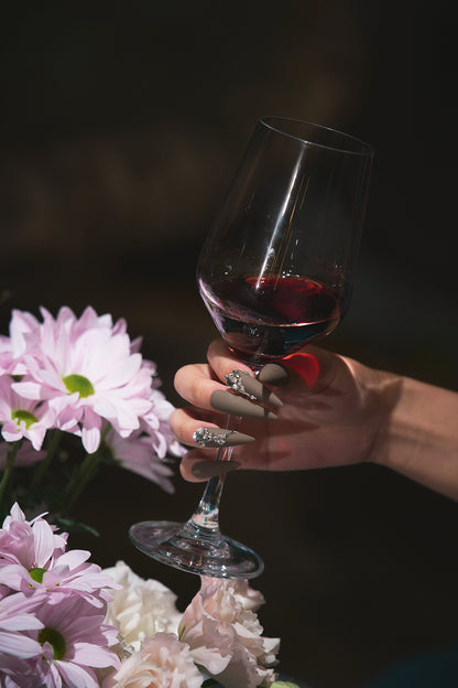 Hand with long brown rhinestone nails holding a glass of red wine, with soft pink flowers in the foreground.