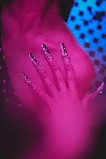 Hand wearing MiuNail Divine Plea black and white cross press-on nails resting on chest, under dramatic purple and blue lighting with soft polka dot elements.