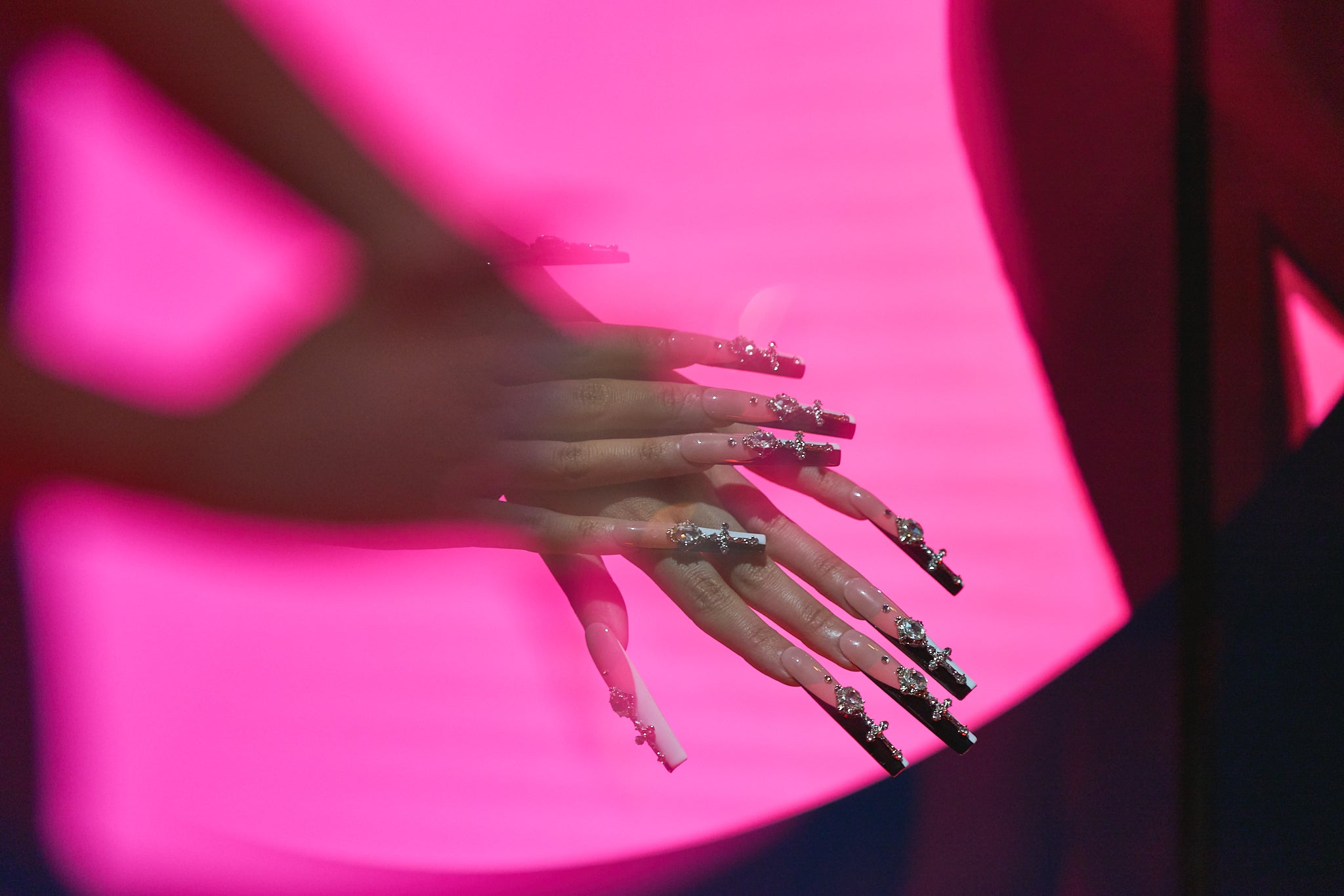 Two hands featuring MiuNail Divine Plea cross press-on nails layered in front of a vivid pink background, showcasing high-shine rhinestones and black-white contrast tips.