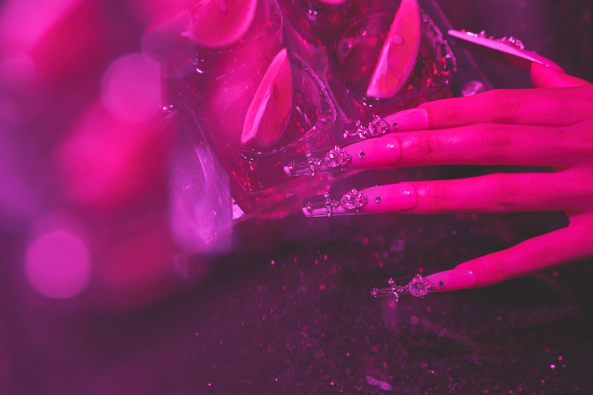 Close-up of a hand wearing Divine Plea press-on nails by MiuNail, featuring gothic crosses and rhinestones, reaching toward tequila shots with lime wedges under moody pink lighting.