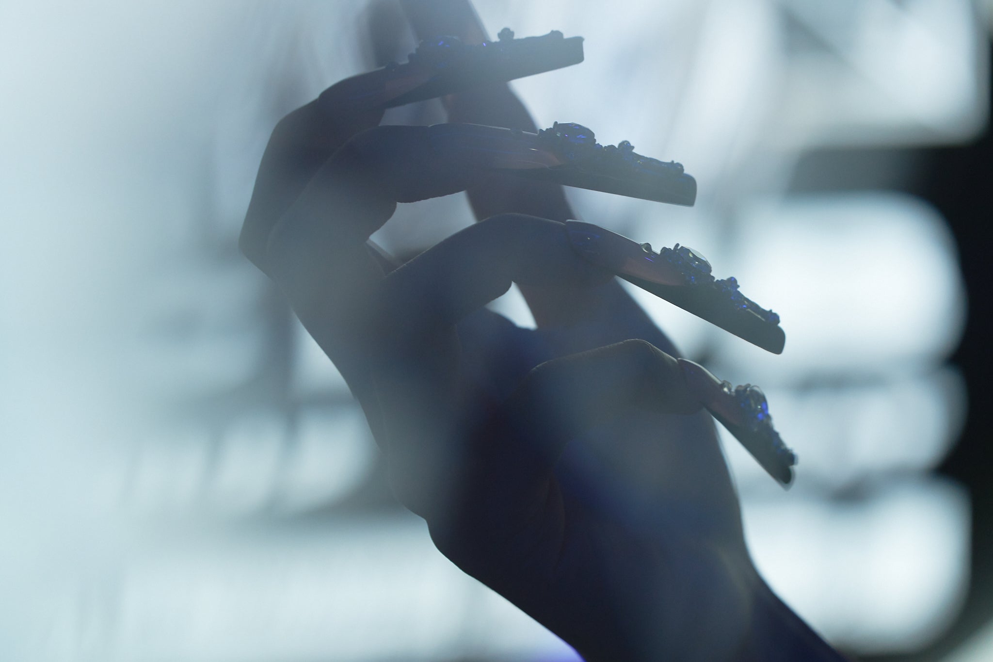 Silhouette of a hand wearing Divine Plea press-on nails by MiuNail, featuring gothic cross embellishments against a blurred monochrome background.